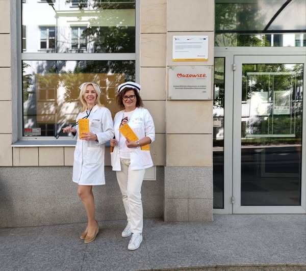 Dwie bibliotekarki pozują przed drzwiami do biblioteki w strojach pielęgniarskich trzymając recepty na książki biblioterapeutyczne w dłoniach