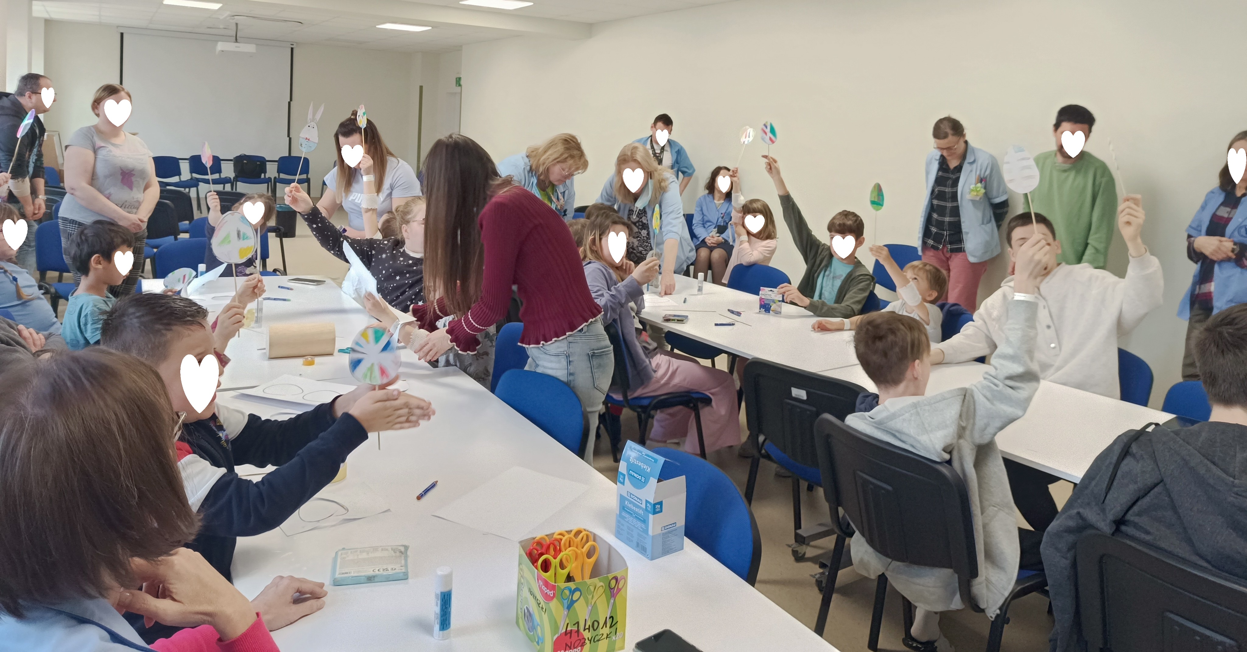 Dzieci pracują przy stolikach z pomocą dorosłych opiekunów tworząc zajączki i pisanki za pomocą kredek, nożyczek, kleju, papieru i patyczków